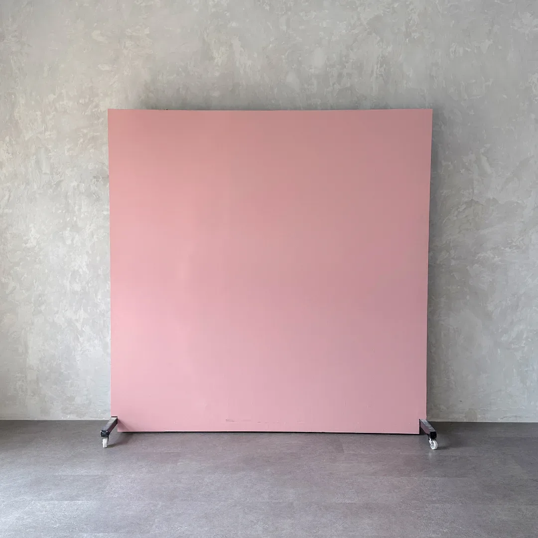 Papan backdrop portable warna pink dusty berdiri di atas roda dan disandarkan ke dinding semen abu bertekstur.