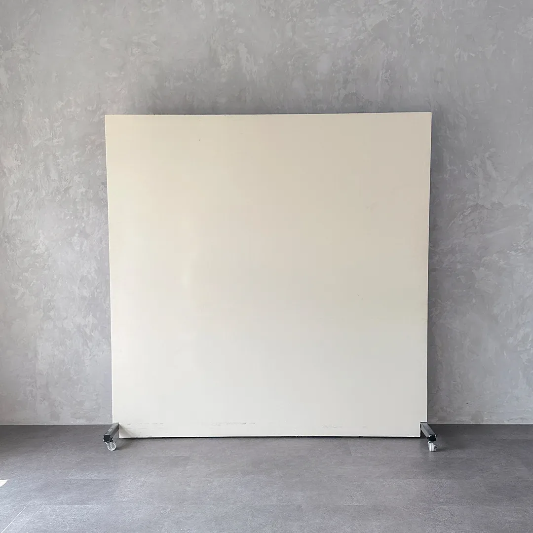 Papan backdrop portable warna krem off-white berdiri di atas roda dan disandarkan ke dinding semen abu bertekstur.