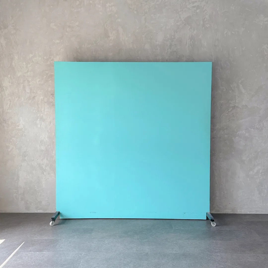 Backdrop portable polos warna biru tosca berdiri di atas roda, disandarkan ke dinding plester abu dengan lantai beton.