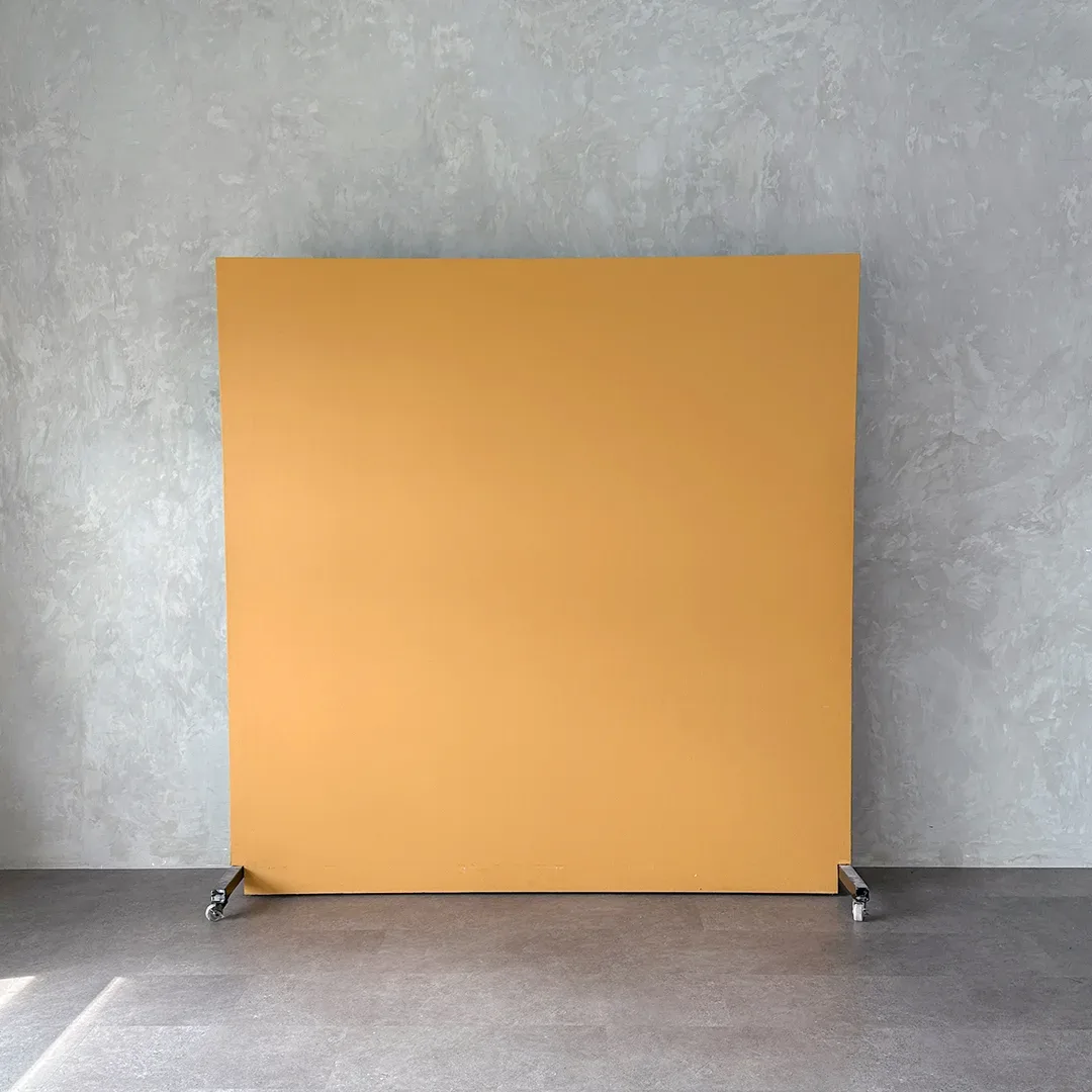 Backdrop portable polos warna kuning mustard berdiri di atas roda dan disandarkan ke dinding plester abu bertekstur.