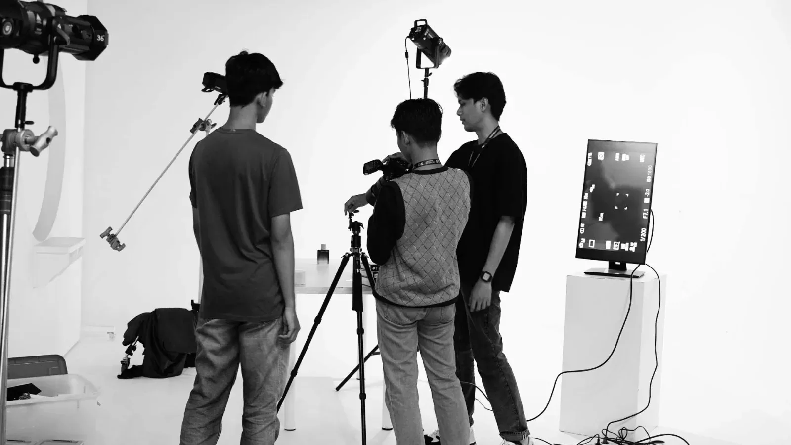 Foto hitam putih tiga kru muda bekerja di studio putih: satu memegang stand, satu di balik kamera pada tripod, dan satu mengawasi monitor tethering di atas box putih.
