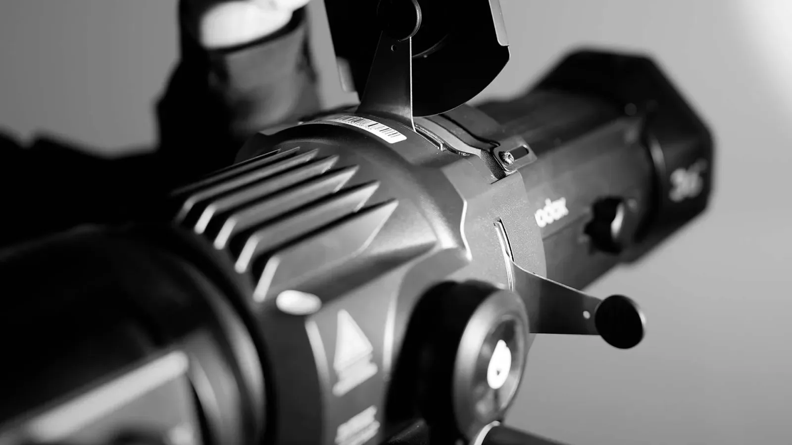Close up hitam putih kepala lampu spotlight fresnel merek Godox lengkap dengan tuas fokus dan sirip pendingin, dengan latar abu-abu bergradasi.
