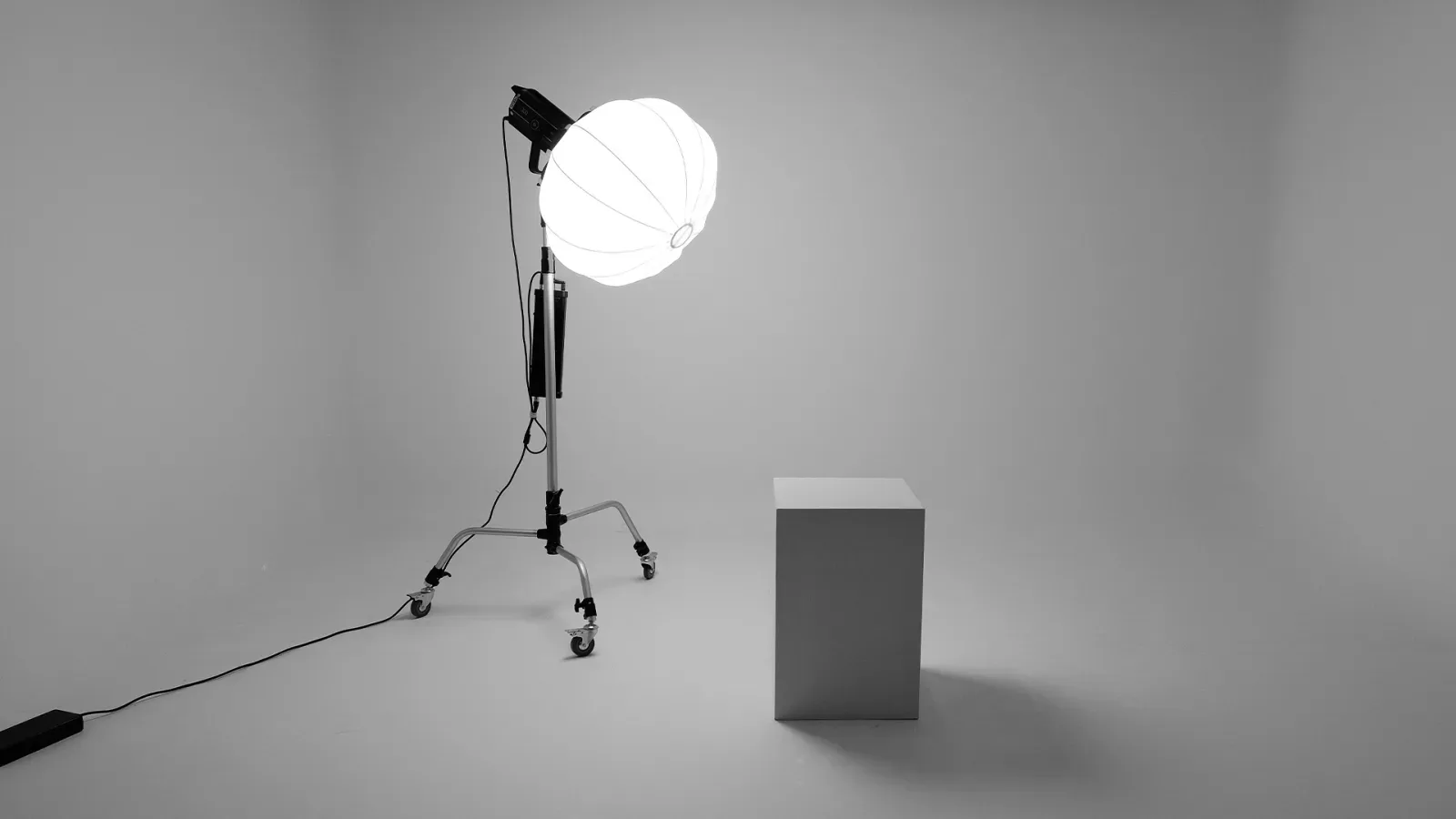 Foto hitam putih ruang studio kosong bercat putih dengan satu lampu continuous berbentuk lantern ball menyala di atas roller stand dan kubus putih sebagai properti duduk.